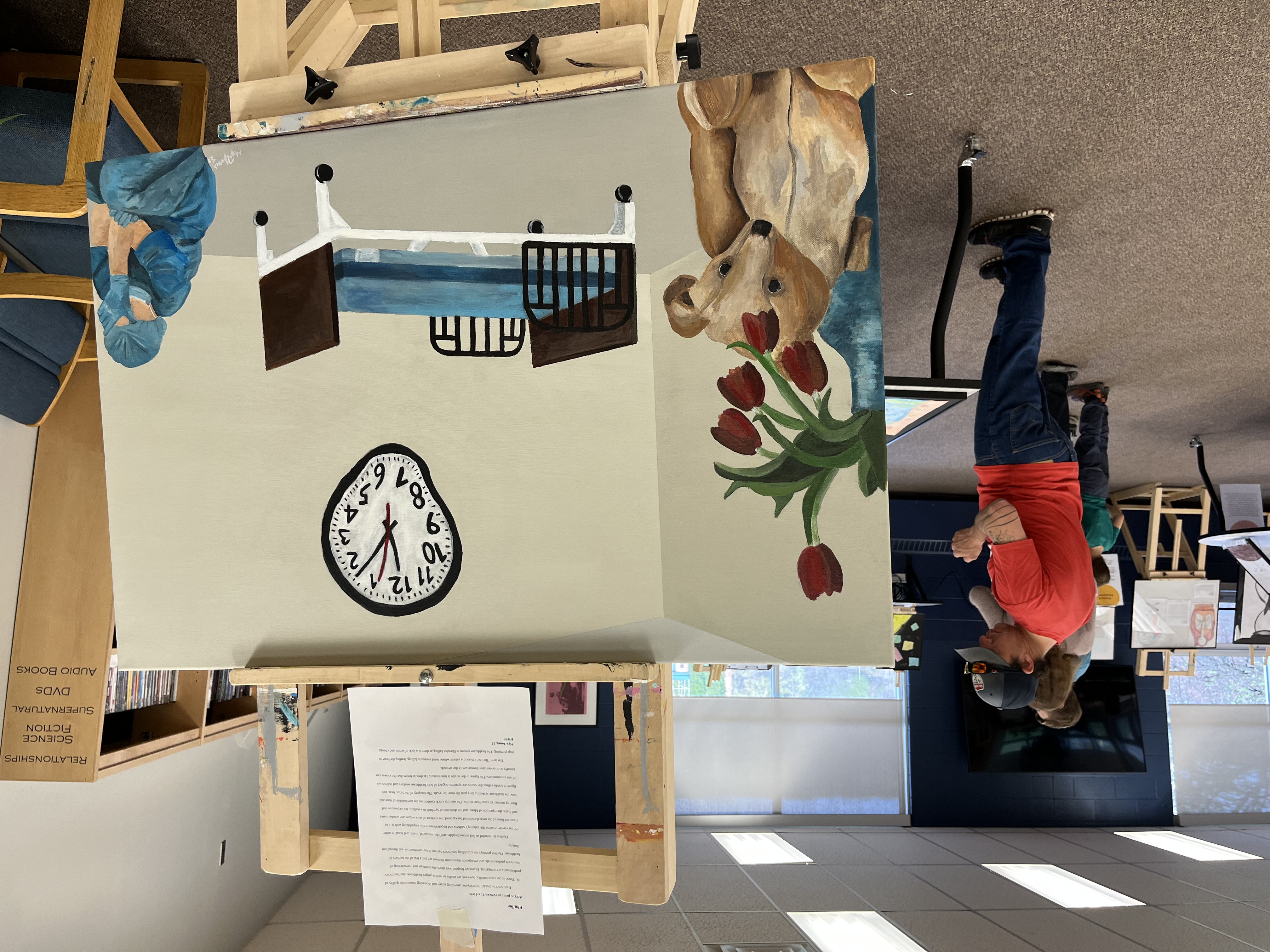 In the foreground, a painting by Mya Jones called Flatline shows an empty hospital bed, a melting clock, wilted tulips and a stuffed animal, and a healthcare worker sitting on the floor with head in hands in despair. In the background, people look at the other pieces of artwork on display.