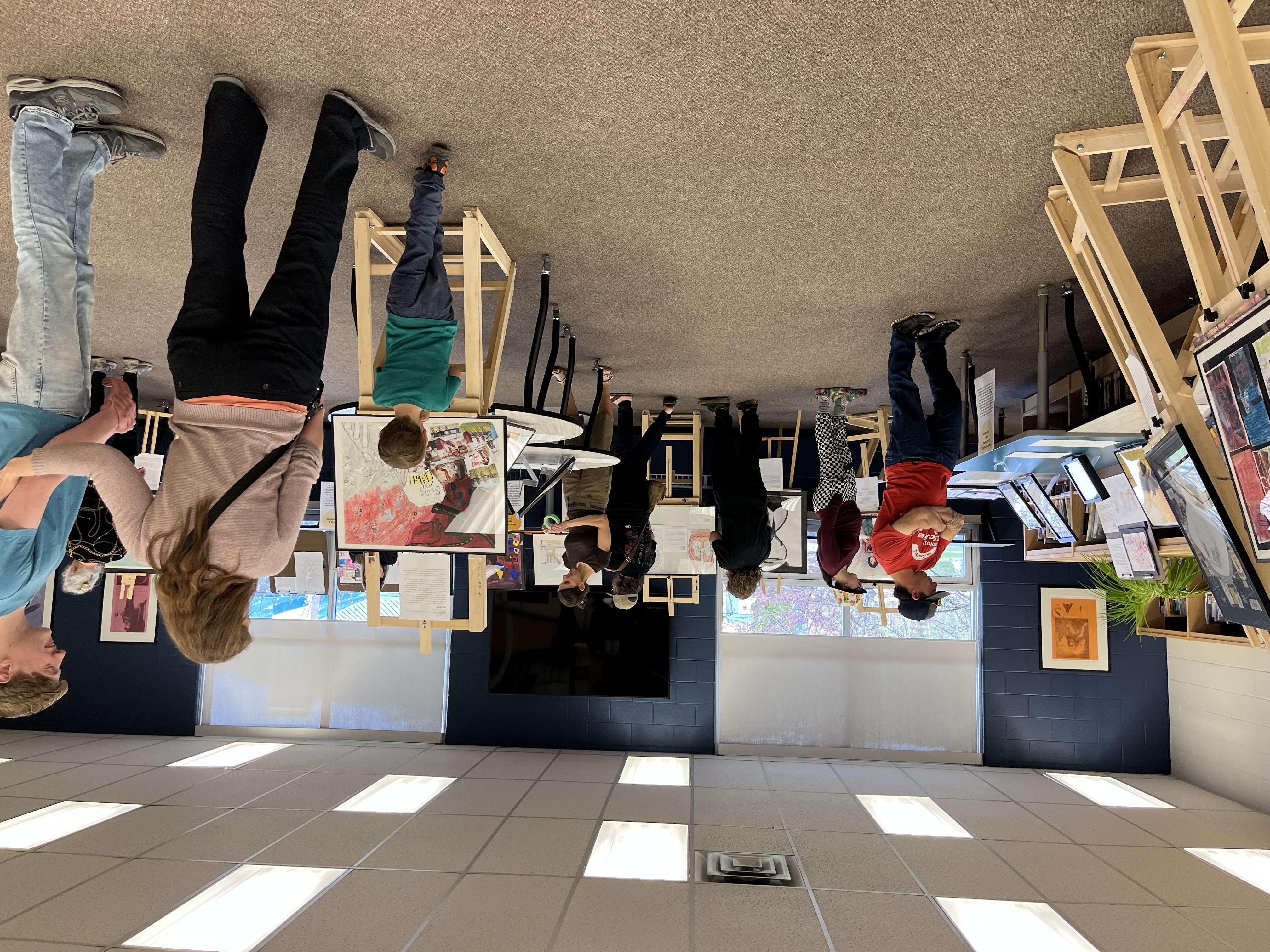 A view of the CSWB Youth Art Exhibition held at HHSS on May 10. Artwork arranged on easels can be seen in foreground and background. People are looking at the art around the room.