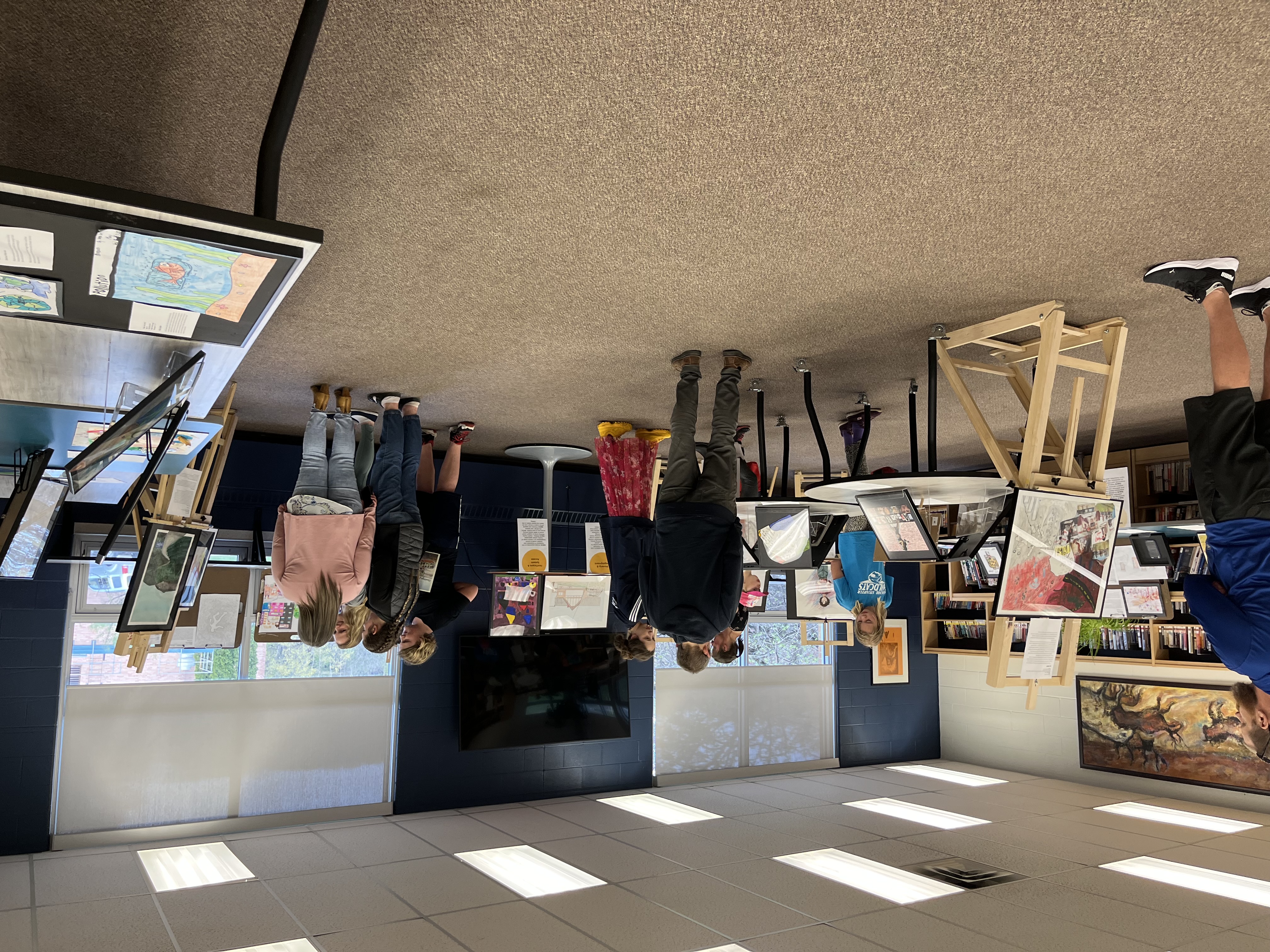 A view of the CSWB Youth Art Exhibition held at HHSS on May 10. Artwork arranged on easels can be seen in foreground and background. People around the room, both kids and adults, are looking at the artwork.