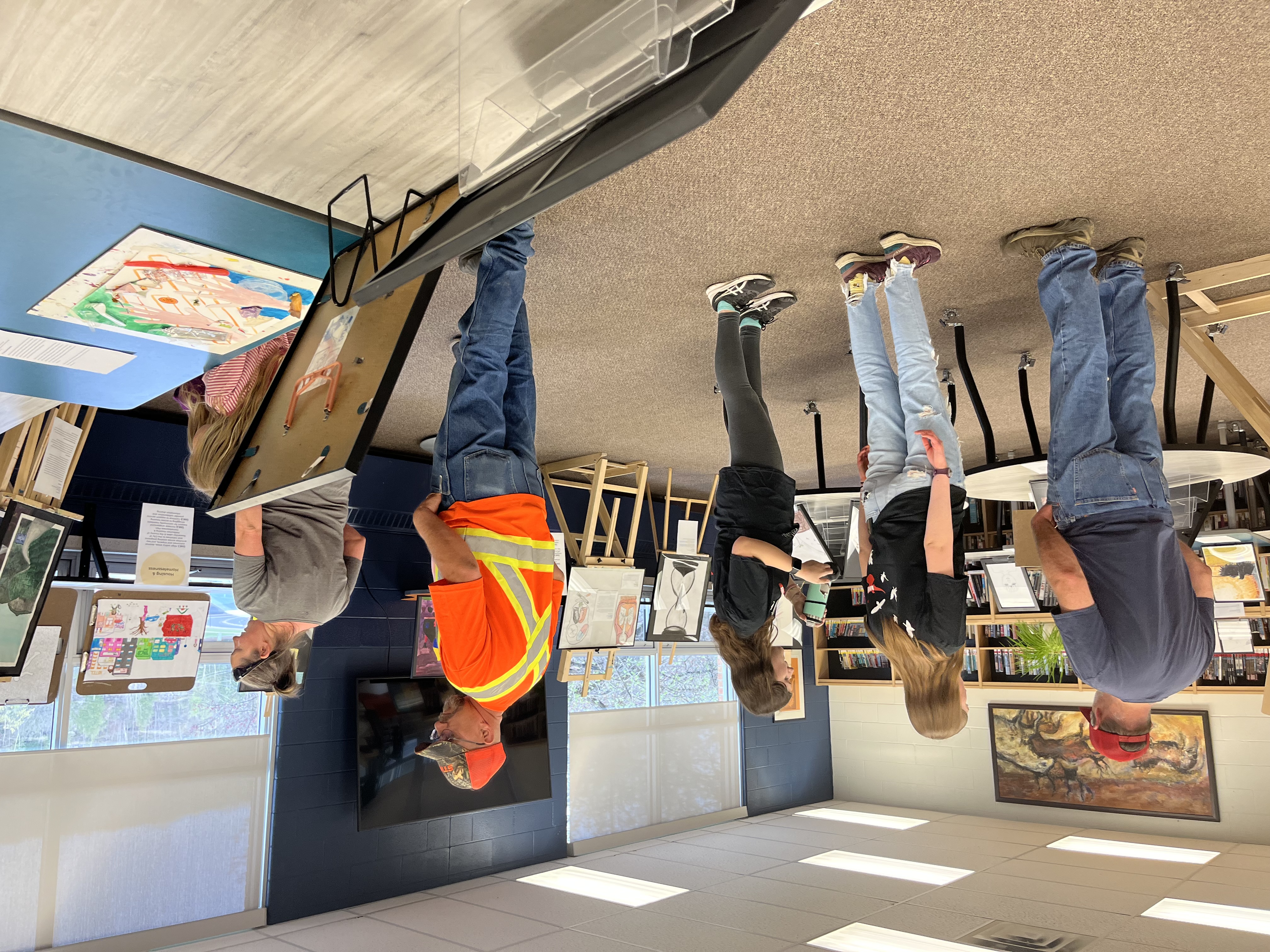 A view of the CSWB Youth Art Exhibition held at HHSS on May 10. Artwork arranged on easels can be seen in foreground and background. People around the room are looking at the artwork. 