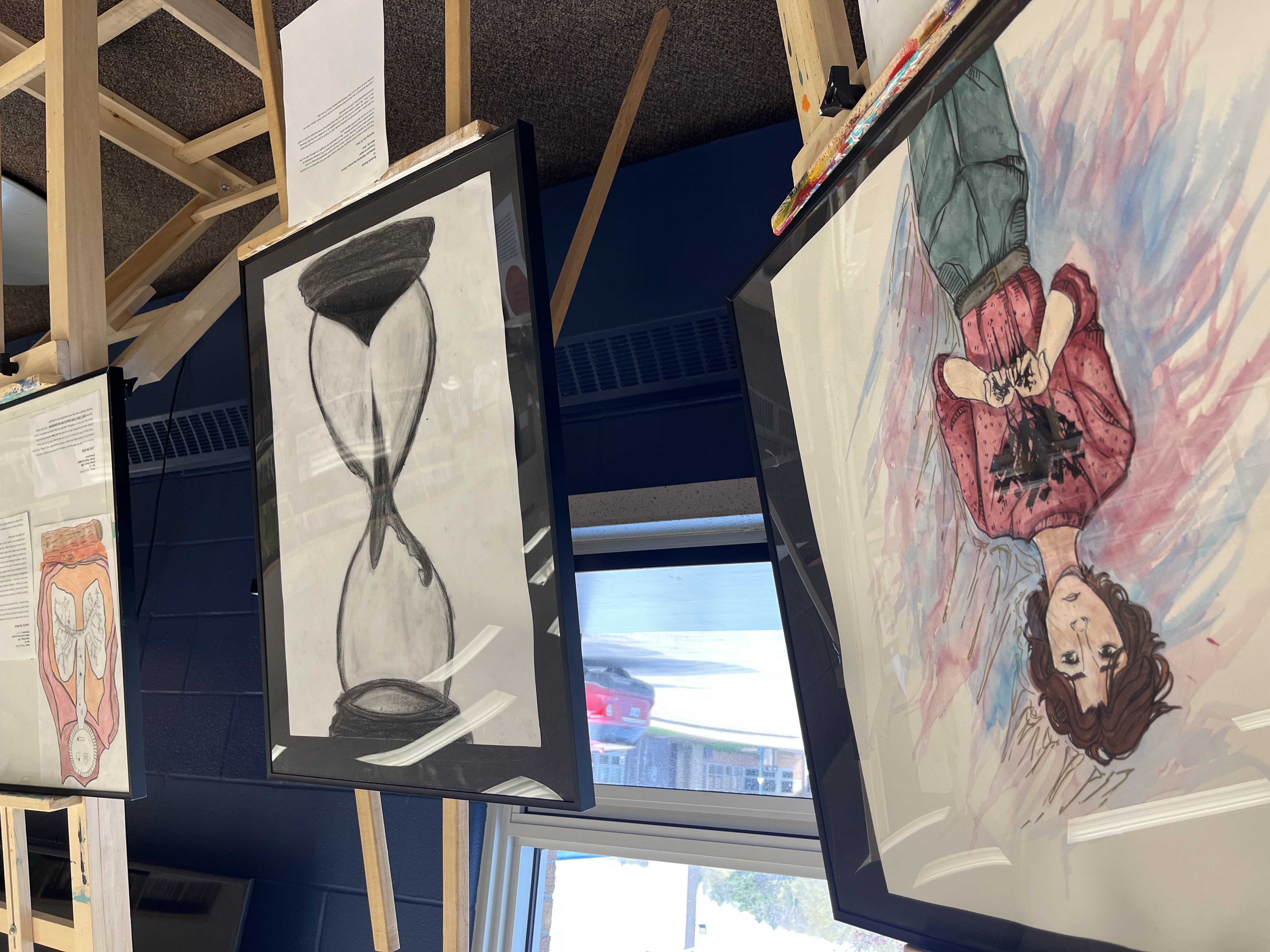 Two art pieces in the CSWB Youth Art Exhibition held at HHSS on May 10 are seen in the foreground. One shows a transgender teen, and one shows a person sitting in the top of an hourglass while their body sifts into the bottom. 